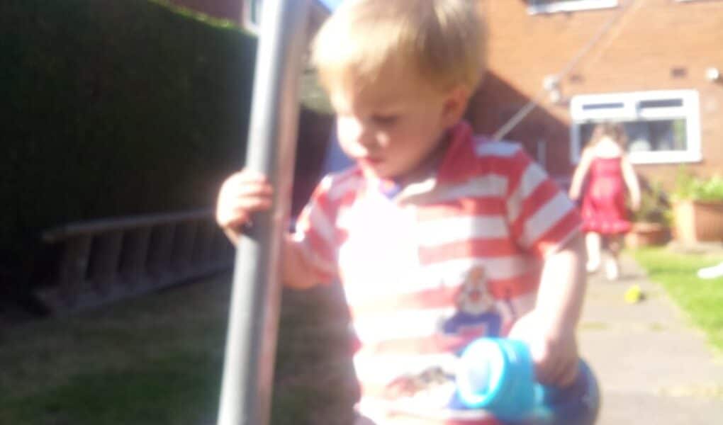 A young child with blonde hair, wearing a red and white striped shirt, holds a blue beaker and stands outdoors near a metal pole. The background is blurry, but another child in a red dress is visible near a brick house and garden.