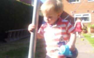 A young child with blonde hair, wearing a red and white striped shirt, holds a blue beaker and stands outdoors near a metal pole. The background is blurry, but another child in a red dress is visible near a brick house and garden.