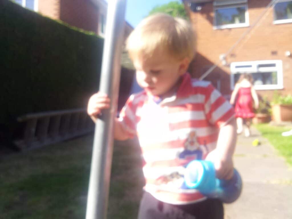 A young blond child in a red-and-white striped shirt holds a blue bottle and stands near a pole outdoors. The background is blurred but shows another child in a red dress, grass, and a brick house with windows. It is a sunny day.