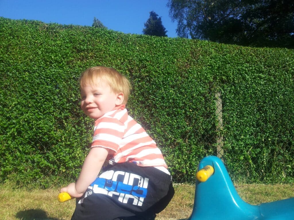 A young child with short blond hair, wearing a red and white striped shirt and dark shorts, sits on a blue and yellow playground rocker in a sunny park. Behind him is a tall green hedge and a wire fence under a clear blue sky.