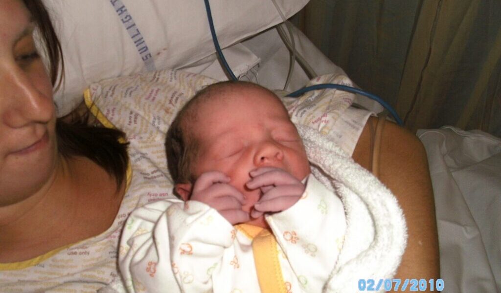 A woman lies in a hospital bed, holding a newborn baby wrapped in a white blanket. The baby’s eyes are closed and its hands are near its face. The woman’s face is partially visible. The date “02/07/2010” is printed in blue on the photo.