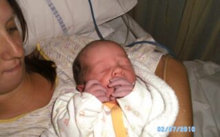 A woman lies in a hospital bed, holding a newborn baby wrapped in a white blanket. The baby’s eyes are closed and its hands are near its face. The woman’s face is partially visible. The date “02/07/2010” is printed in blue on the photo.
