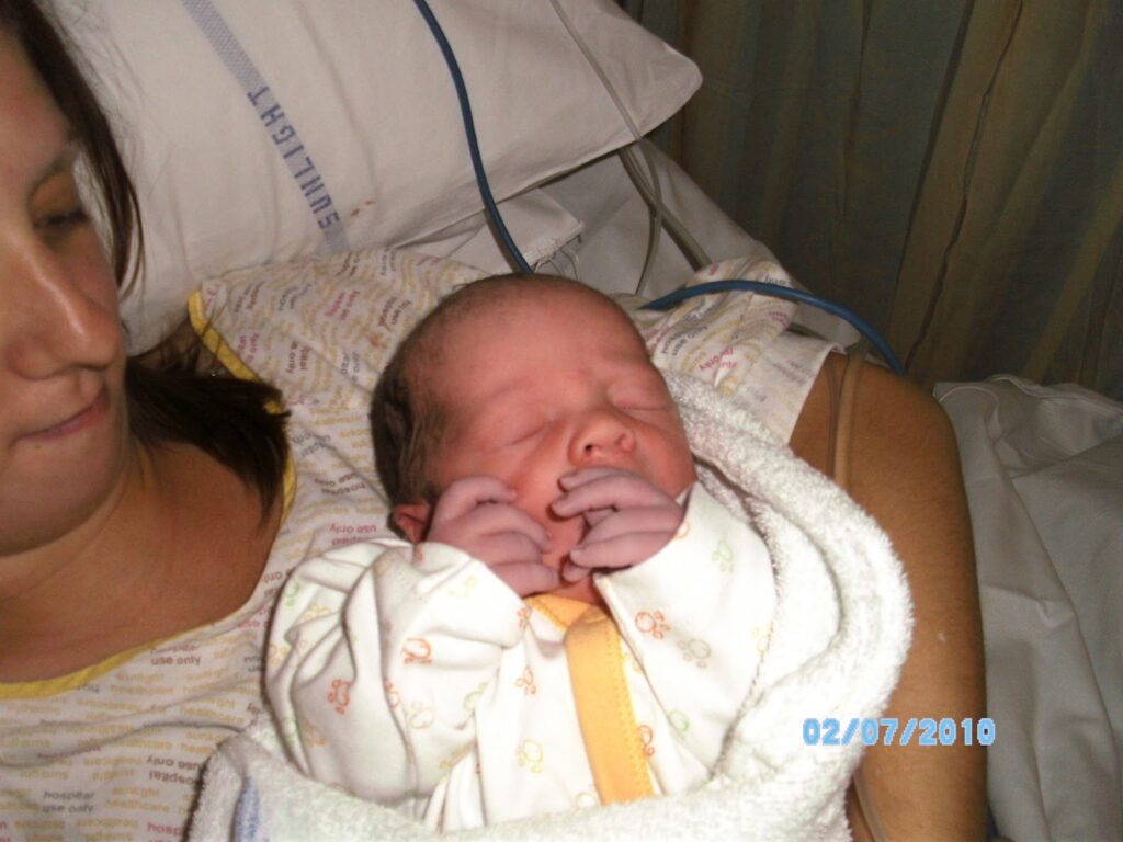 A woman in a hospital gown holds a newborn baby wrapped in a white blanket with yellow patterns. The baby’s hands are near its face. Medical equipment and bedding are visible in the background. The date “02/07/2010” is displayed in blue text.