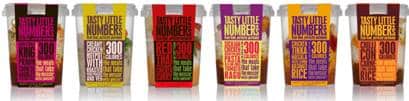 Six colourful pots of "Just Little Numbers" snacks are lined up. Each pot has a distinct colour label and bold text highlighting that each serving contains around 300 calories. Flavours are visible but not legible due to the small text.