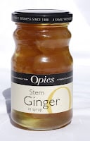 A glass jar of Opies Stem Ginger in Syrup with a black lid. The jar is filled with light golden syrup and pieces of ginger. The label is cream and black, displaying the brand name "Opies" and product name in clear text.