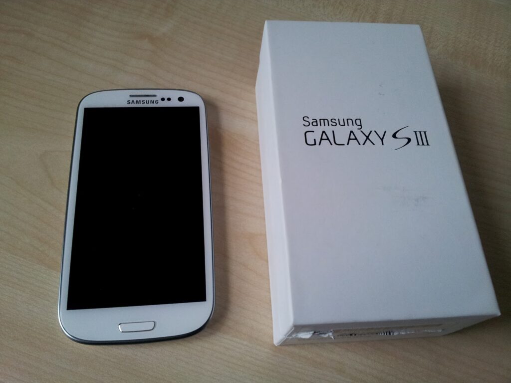 A white Samsung Galaxy S III smartphone lies on a light wooden surface next to its white retail box, which is labelled āSamsung GALAXY S IIIā in black text. The phoneās screen is off, and both objects are seen from above.