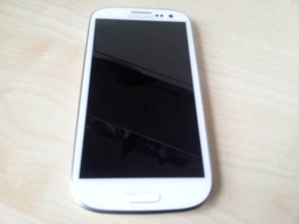 A white Samsung mobile phone with a black screen lies face up on a light-coloured wooden surface. The phone has rounded edges and a physical home button at the bottom centre. The screen is off, reflecting some light and faint shapes.