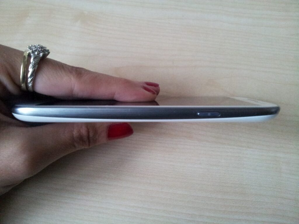 A hand with red nail varnish and a ring holds a mobile phone horizontally, showing a noticeable curve in the phoneās body. The phone appears slightly bent and is viewed from the side against a light wooden background.