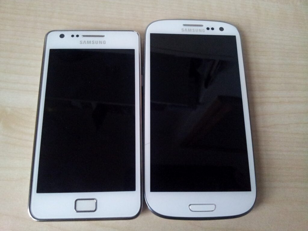 Two white Samsung smartphones are placed side by side on a light wooden surface. The phone on the left is smaller with a square home button, whilst the phone on the right is larger with a rectangular home button. Both screens are black and switched off.
