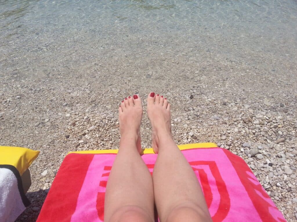 A person with painted red toenails relaxes on a colourful pink and red beach towel, sunbathing by a pebbled shoreline with clear, shallow water in the background. A corner of a yellow and white towel is visible on the left.