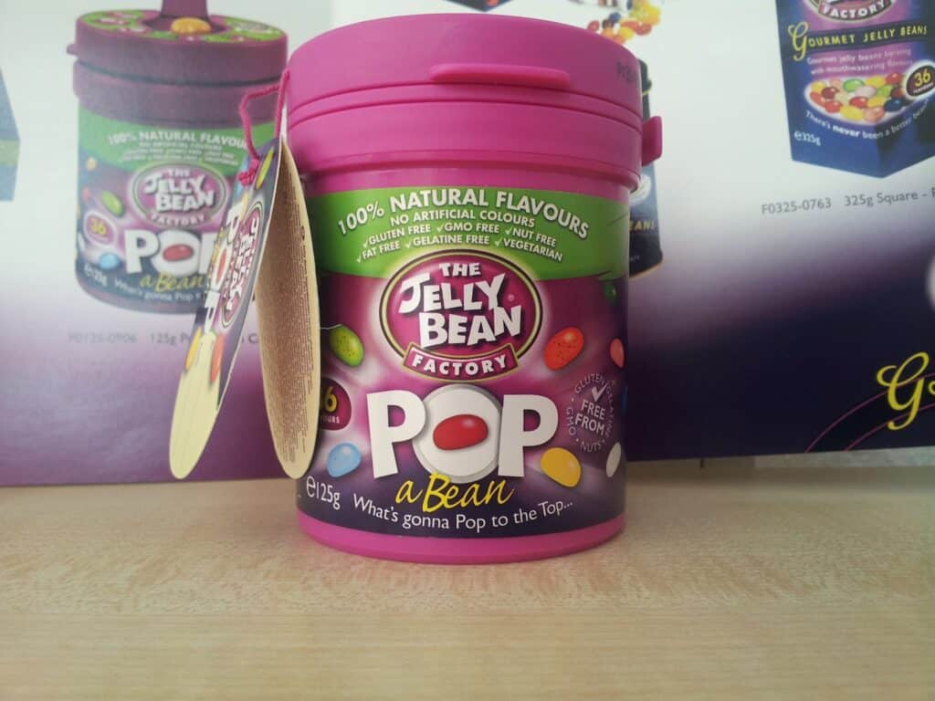 A pink cylindrical tub labelled "The Jelly Bean Factory POP a Bean" sits on a table. The label advertises "100% natural flavours" and includes colourful jelly beans. A product tag hangs on the side, and a display with more sweets is blurred in the background.
