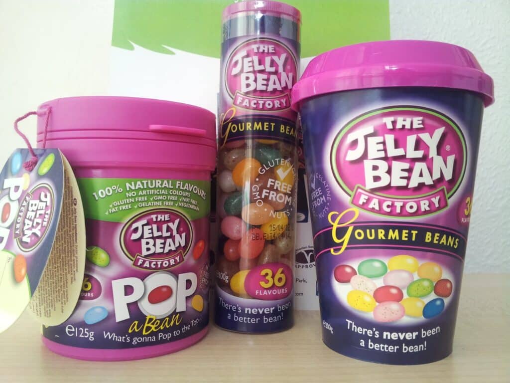 Three colourful containers of The Jelly Bean Factory Gourmet Beans are shown. Each has a purple and pink design with images of jelly beans. One is a tub, one is a tall tube, and one is a lidded cup. Text promotes natural flavours and 36 different varieties.