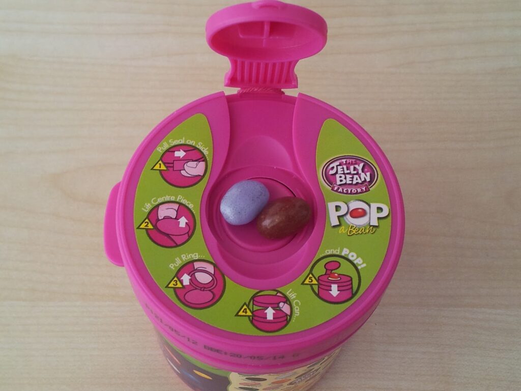 A pink container labelled "The Jelly Bean Factory Pop a Bean" is open, showing two jelly beans inside—one purple, one brown. The lid displays illustrated instructions in yellow circles showing how to open, pull, and pop jelly beans.
