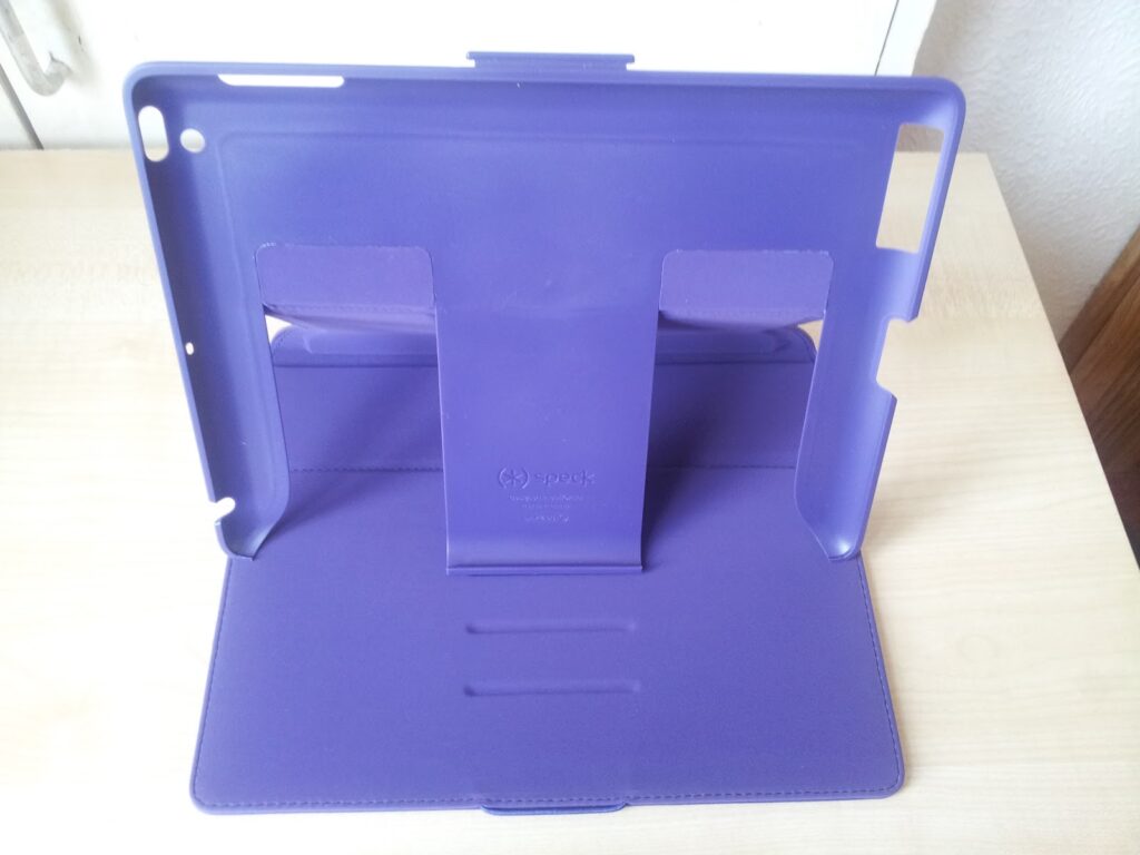 A purple tablet case stands open on a light wooden surface. The case has cut-outs for buttons and ports, two foldable inner flaps for propping up a tablet at different angles, and a smooth back. The setting appears to be indoors near a white wall.