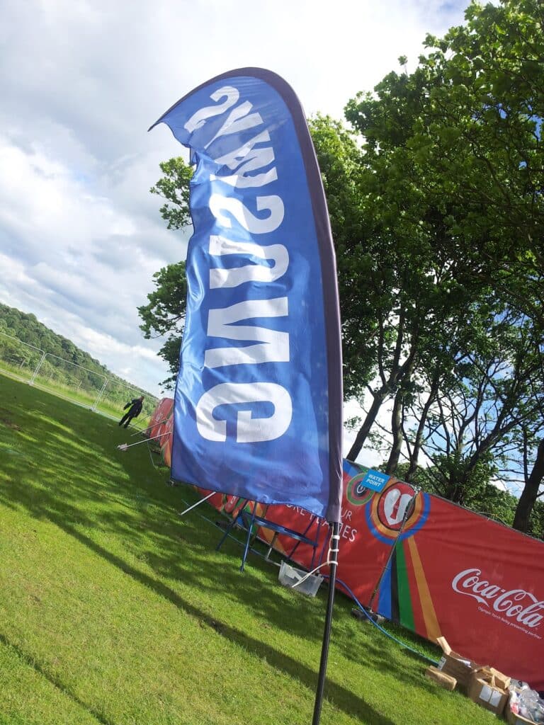 A blue flag with "SAMSUNG" written in white appears reversed due to the angle. It stands on a grassy field near trees and a red Coca-Cola banner. The sky is partly cloudy, and some event equipment is visible in the background.