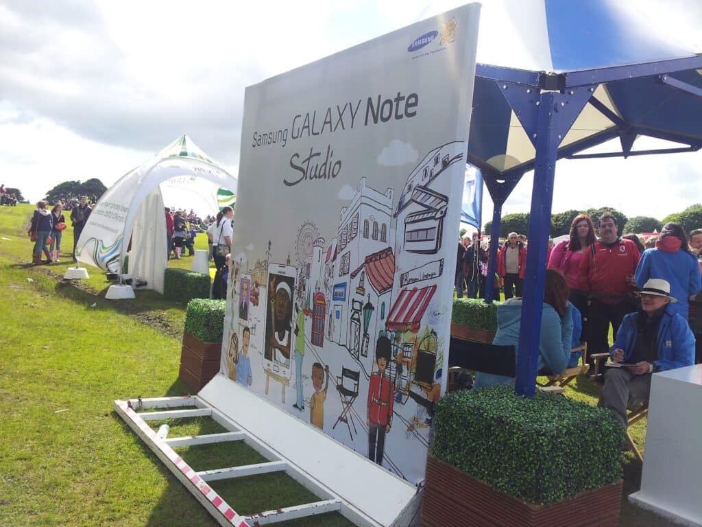 A large Samsung Galaxy Note Studio sign with colourful illustrations stands on grass at an outdoor event. People in colourful jackets gather near tents and stalls, while some sit under a blue and white canopy. The sky is partly cloudy.