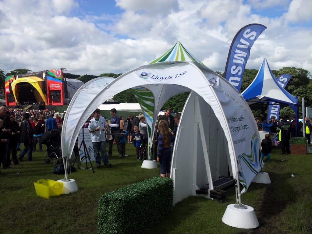 A festival scene shows a crowd near a large stage with colourful screens. In the foreground, there is a white tent with Lloyds TSB branding. People, including children, walk on the grass. Flags, small hedges, and other tents are visible on a partly cloudy day.