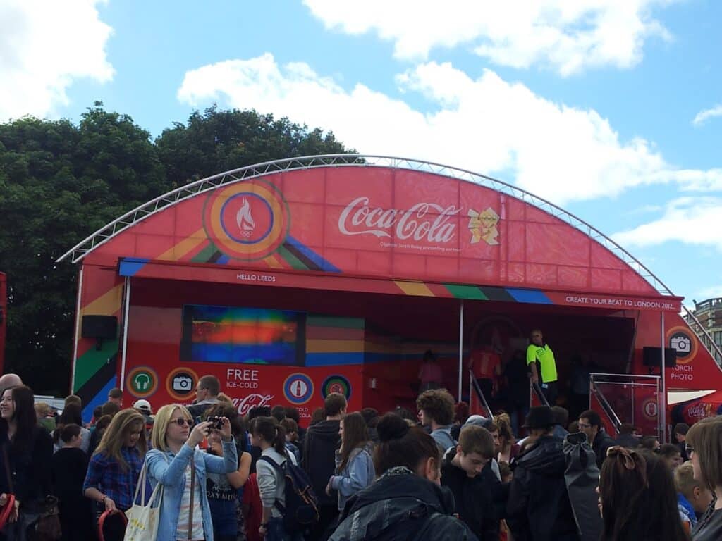 A crowd gathers in front of a bright red Coca-Cola event stand with a curved roof and Olympic branding. People take photos, talk, and queue while staff in neon jackets assist guests. Green trees and buildings are visible in the background under a partly cloudy sky.