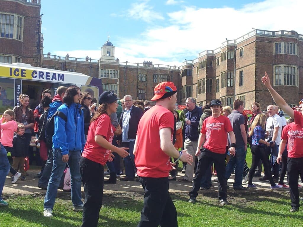 A crowd of people, many in red shirts, gather outdoors near a historic brick building. Some talk and walk, while others stand in groups. An ice cream van is visible on the left. The weather is sunny with blue sky and scattered clouds.