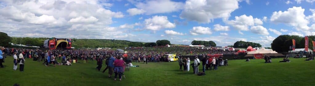 A large outdoor music festival scene with crowds gathered near a stage on the left. People are spread across a green grassy field under a bright sky with scattered clouds. Tents, food stalls, and banners are visible in the background.