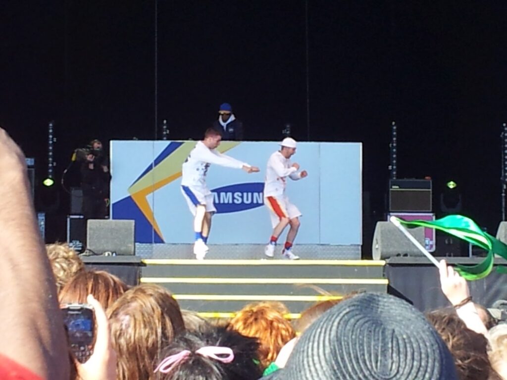 Two men in sportswear break-dance on a stage outdoors, backed by a Samsung sign and a DJ. An audience watches, with some holding up mobile phones or waving a green scarf. The scene is sunny, and the stage is set against a black backdrop.