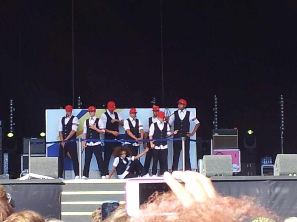 A dance group of eight men in matching black waistcoats, white shirts, black trousers, and red caps perform on an outdoor stage. Seven stand in a line holding canes, while one kneels in front. Members of the audience watch, with one person holding up a mobile to record.