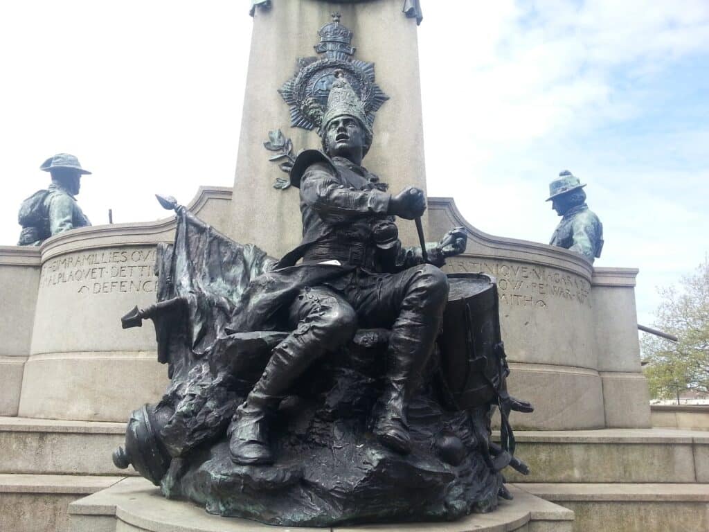 A bronze statue of a seated soldier holding a flag is in front of a tall stone war memorial. Two other soldier statues in different uniforms stand behind on either side. Inscribed plaques and detailed reliefs are visible on the memorial, with a partly cloudy sky above.