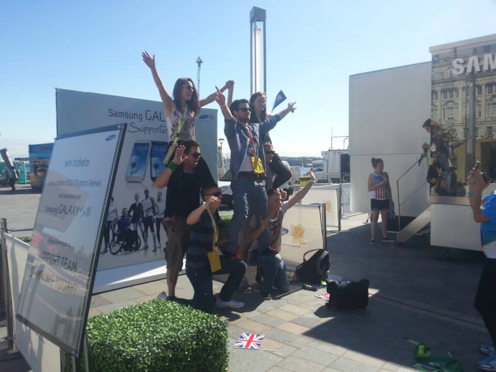 A group of seven people form a cheerleader-style human pyramid outdoors, smiling and raising their arms. Behind them are Samsung Galaxy promotional banners. The sun is bright, and some small flags are on the pavement nearby. Other people watch the scene.