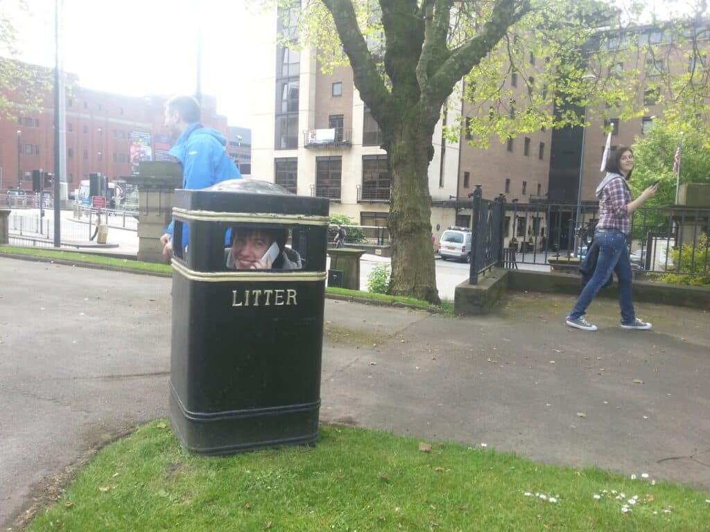 A man sits inside a large black rubbish bin with his head poking out of the opening, smiling. Another man in a blue jacket stands nearby on the pavement, and a woman in a checked shirt walks by, glancing at the scene. Trees and buildings are in the background.