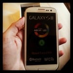A hand holds a white Samsung Galaxy S III smartphone. The screen displays the startup logo with “GALAXY S III” at the top and an Android logo in the centre. The background is dark, and a phone box is blurred in the background.