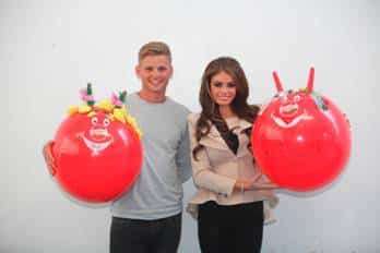 A smiling young man and woman stand side by side, each holding a large red balloon decorated with cartoon faces and colourful features like horns and flowers. They are in front of a plain white wall. Both look cheerful and are dressed casually.