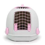 A round, white pet carrier with pink accents and a black metal front door. The door has a grid pattern and two pink catches, and there are ventilation holes at the top. The carrier is designed for small animals like cats or small dogs.