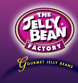 A purple logo features "The Jelly Bean Factory" in bold white letters inside a pink oval. Below, a blue ribbon reads "Gourmet Jelly Beans" in yellow, with a large script "G." The background fades from dark to light purple.