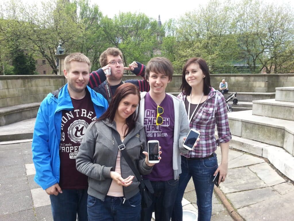 Five young adults stand outdoors by stone steps and curved seating, smiling at the camera. Three of them hold mobile phones, one presenting hers up close. Trees and a statue are in the background; it’s a bright day, and everyone is casually dressed.