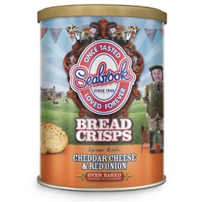 A cylindrical tin of Seabrook Bread Crisps is shown. The label features a pastoral scene with cows, a man in tweed, and festive bunting. The flavour is "Cheddar Cheese & Red Onion." The Seabrook logo and slogans are prominently displayed on the front.