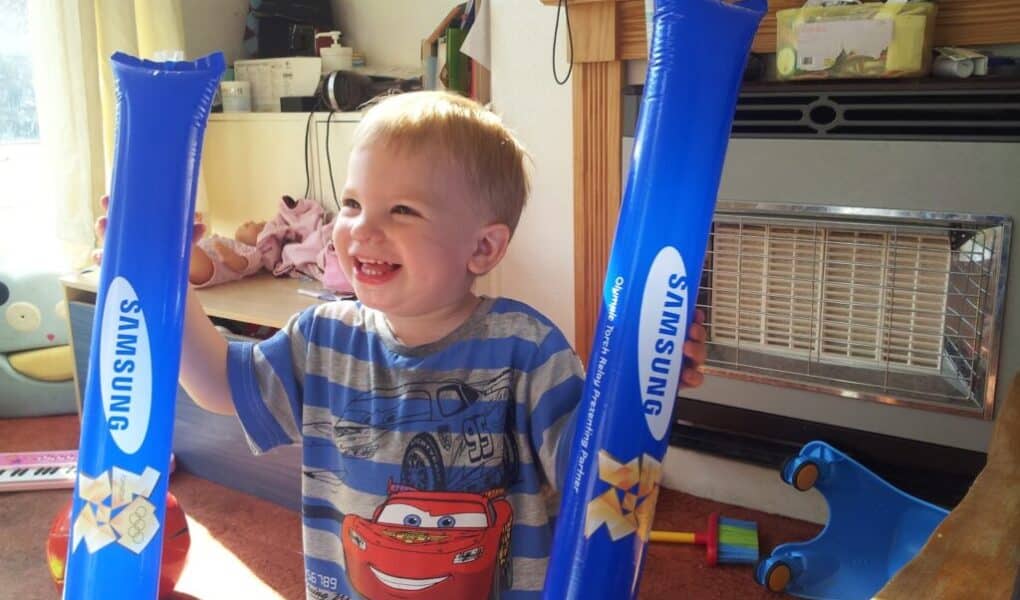 A young child smiles widely indoors, holding two blue inflatable sticks with "SAMSUNG" printed on them. He wears a striped shirt featuring Lightning McQueen from Cars. Sunlight streams through a window; toys and household items are visible in the background.