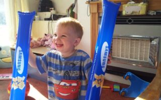 A young child smiles widely indoors, holding two blue inflatable sticks with "SAMSUNG" printed on them. He wears a striped shirt featuring Lightning McQueen from Cars. Sunlight streams through a window; toys and household items are visible in the background.