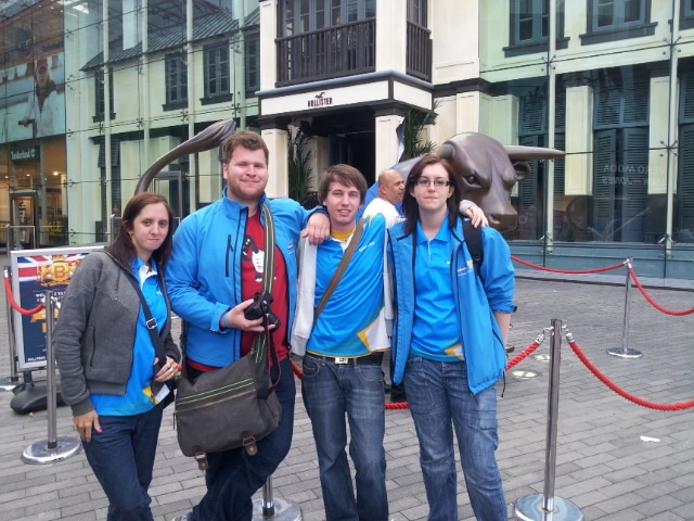 Five people stand together outside, all wearing blue shirts and jackets. Four pose with arms round each other, smiling, whilst one holds a camera. Behind them is a bull sculpture, a red rope barrier, and modern glass-fronted buildings.