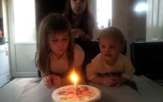 Three young children gather around a birthday cake with a single lit candle. Two girls, one in front and one standing behind, are blowing at the candle. A small boy sits beside the cake, watching the flame. Sunlight filters through a window in the background.