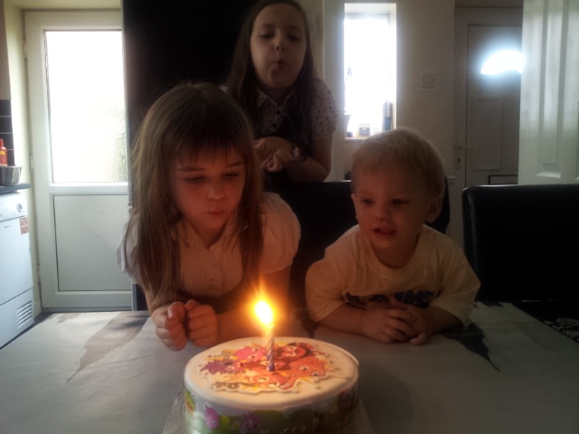 Three young children gather around a birthday cake with a single lit candle. Two girls, one in front and one standing behind, are blowing at the candle. A small boy sits beside the cake, watching the flame. Sunlight filters through a window in the background.