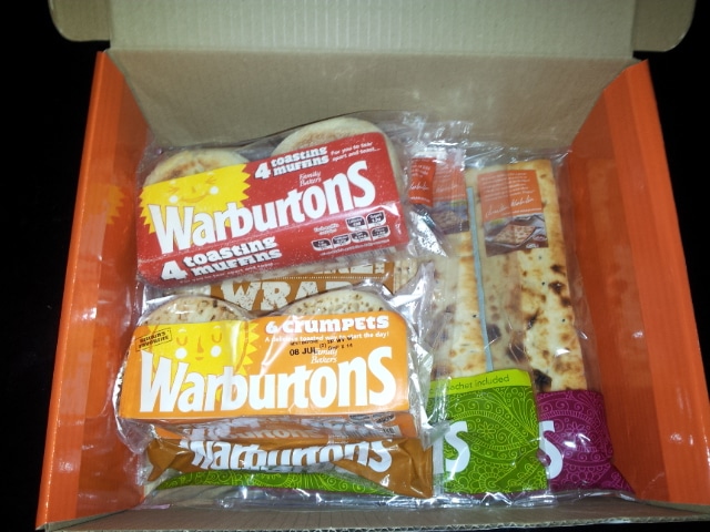 An open cardboard box contains Warburtons branded baked goods in plastic packaging: two packs of four toasting muffins (red and white packaging) and one pack of six crumpets (orange and yellow). Two wrapped flatbreads are also visible.