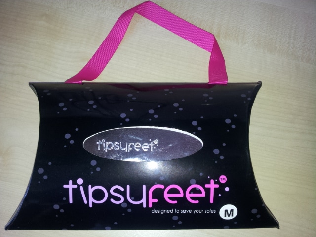 A black pillow-shaped box with white and pink text reading "tipsureet" and “designed to save your soles.” The box has small white dots and a bright pink carrying handle. A silver oval label displays the brand name. A white “M” indicates the size.