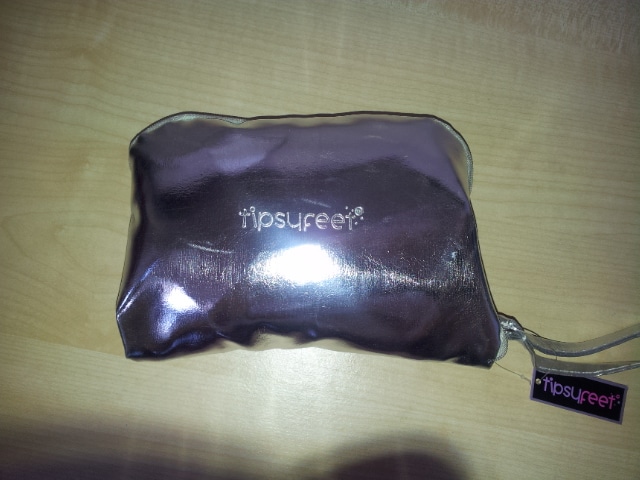 A small, shiny, metallic silver pouch lies on a light wooden surface. The pouch features the brand name "tipsyfeet" in white lettering on the front and a matching black label with the same logo attached to the zip.