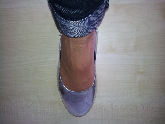 A person’s right foot wearing a light purple or silver flat shoe is shown from above. The shoe has a rounded toe, and the person is wearing dark denim trousers with the cuff turned up slightly. The background is a light wood floor.
