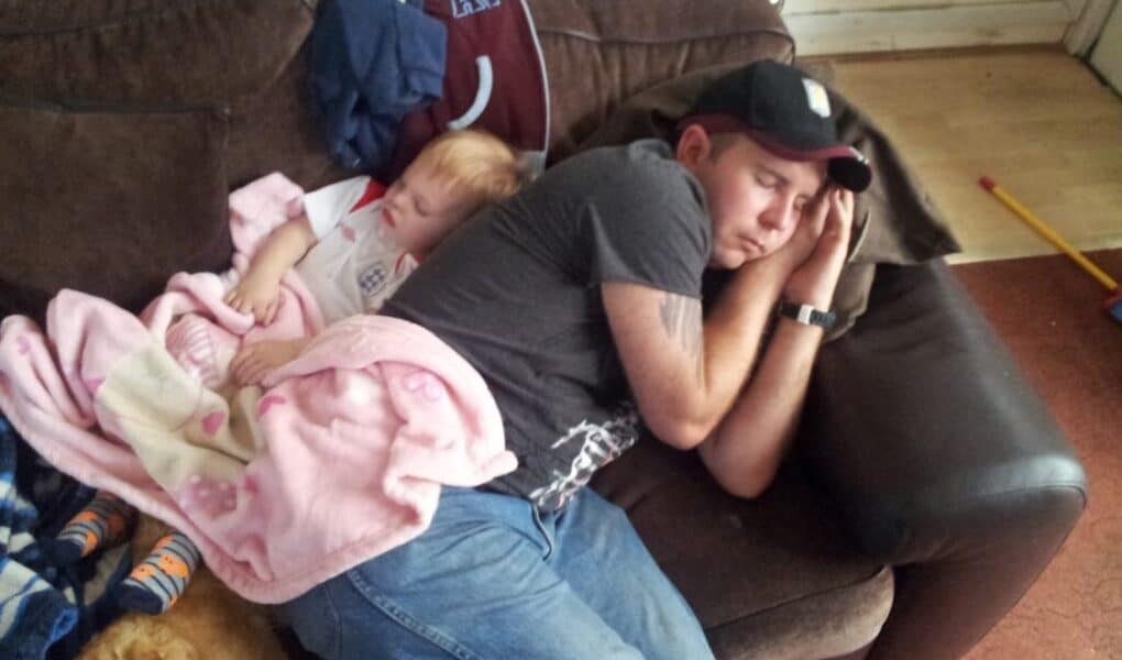 A man wearing a black cap, grey shirt, and jeans naps on a brown sofa, arm under his head. A small child in a white shirt sleeps next to him, wrapped in a pink blanket. A ginger cat lies at their feet. Clothes and toys are visible in the background.