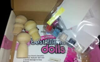 A kit box contains unfinished wooden kokeshi doll figures, small paint pots in various colours, paintbrushes, and paper sheets. The packaging reads “Japanese best friend dolls” across a pink and white background inside the box.