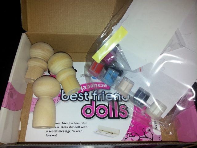 A kit box contains unfinished wooden kokeshi doll figures, small paint pots in various colours, paintbrushes, and paper sheets. The packaging reads “Japanese best friend dolls” across a pink and white background inside the box.