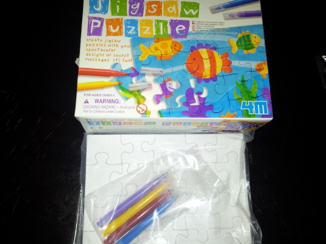 A box labelled "Jigsaw Puzzle" with colourful fish and sea creatures on the cover sits above a blank puzzle and a plastic bag containing four coloured markers. The kit is designed for children to create their own jigsaw puzzles.
