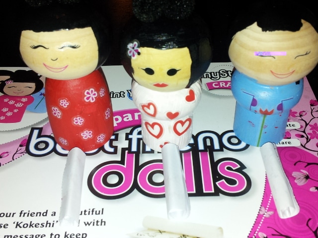 Three wooden Kokeshi-style dolls with painted smiles stand upright. Each wears a different coloured outfit: red with flowers, white with hearts, and blue with fans. They are placed on a card with the words “best friend dolls” in bold text.