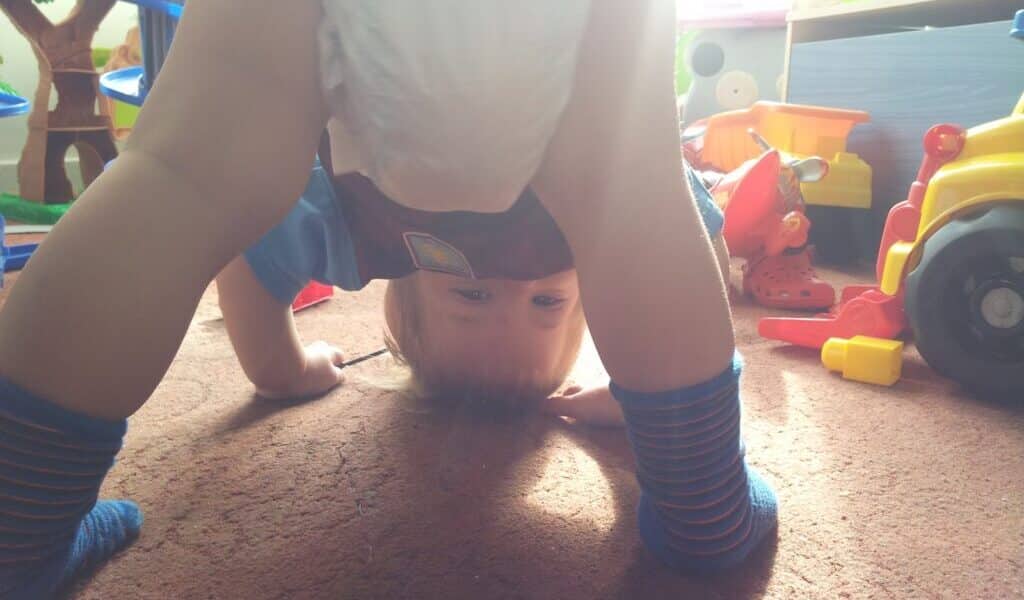 A toddler wearing a nappy and striped socks bends over, looking upside down between their legs in a playroom filled with colourful toys and sunlight streaming in from behind.